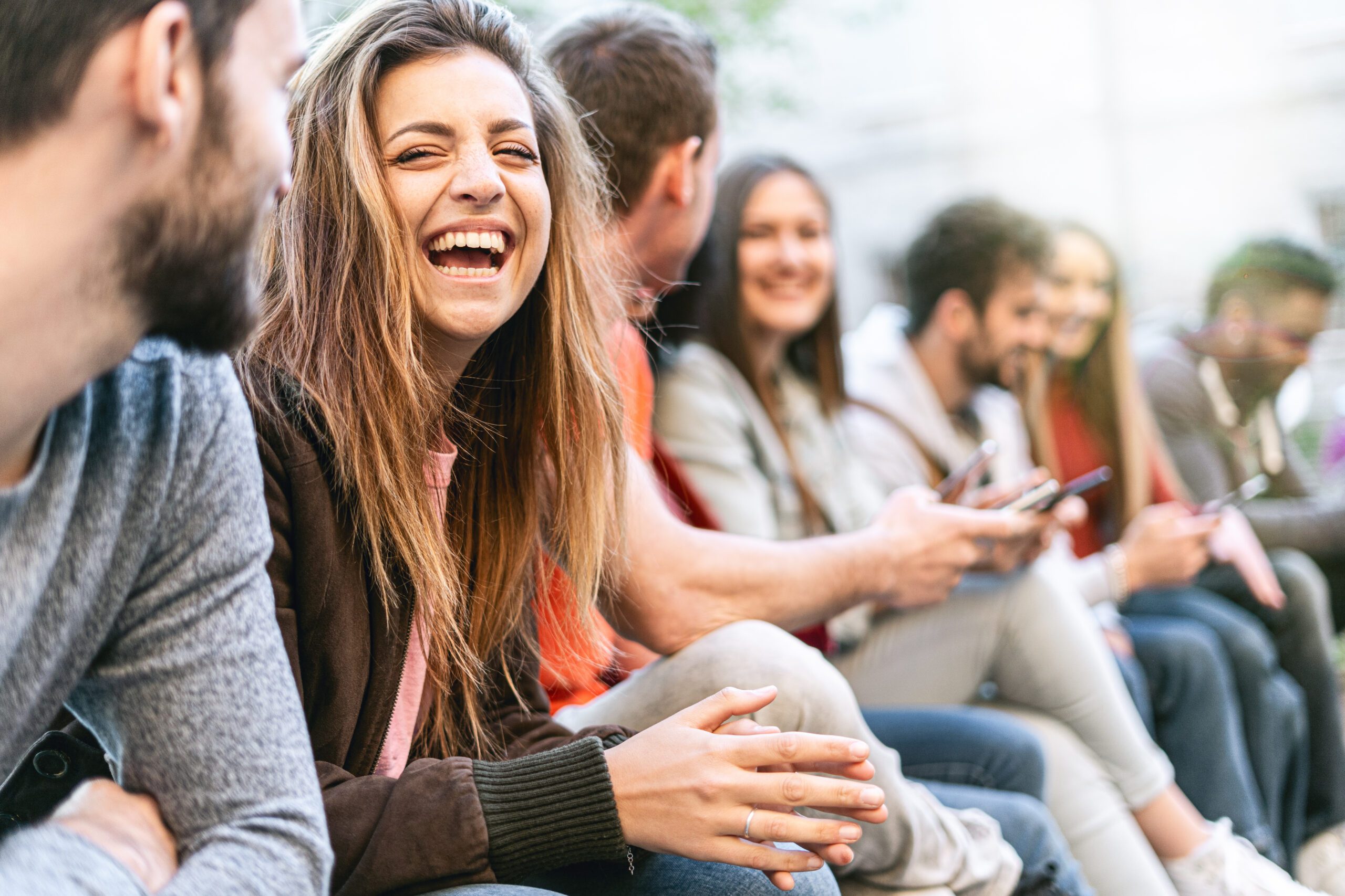 Group of trendy young people chatting together sitting on a bench outdoors. Students having fun together. Focus on a blonde girl smiling with open mouth